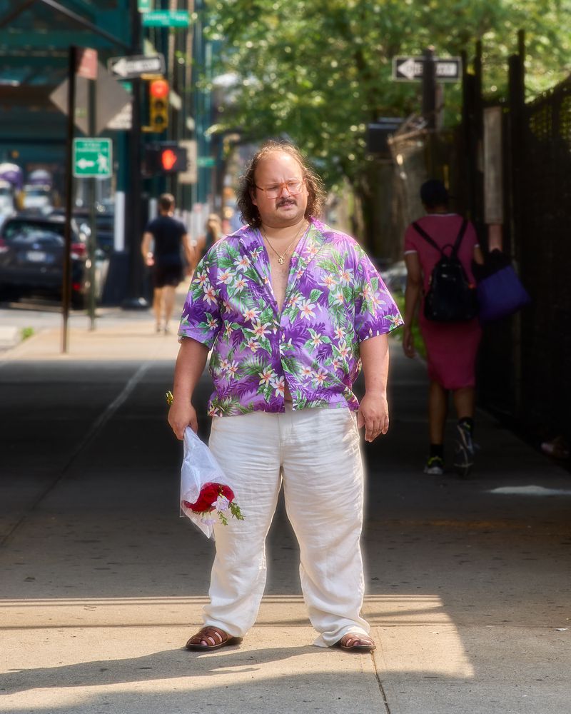 Man stands on a city sidewalk wearing a Hawaiian shirt, white pants, and sandals while holding a bouquet of red and white flowers.