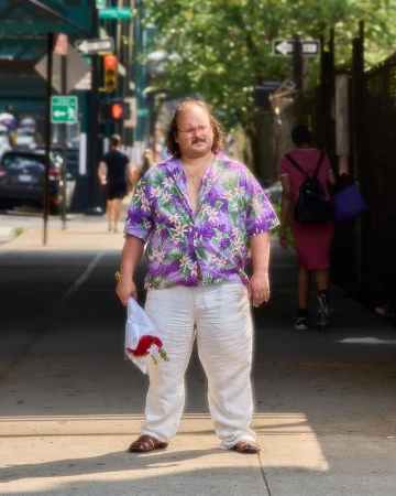 Man stands on a city sidewalk wearing a Hawaiian shirt, white pants, and sandals while holding a bouquet of red and white flowers.