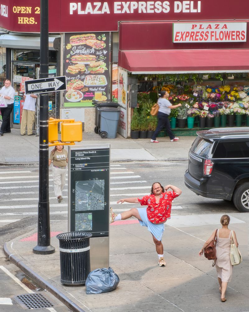 Man in red shirt does high kick on city street with flower shop, deli, and pedestrians visible. Travel, urban scene, daytime.