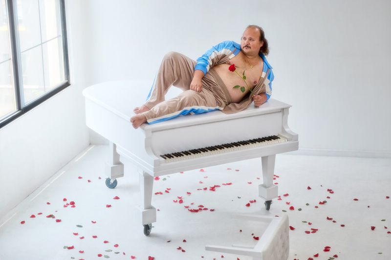 Adult male reclining on a white grand piano in a white room decorated with rose petals. He is holding a red rose.