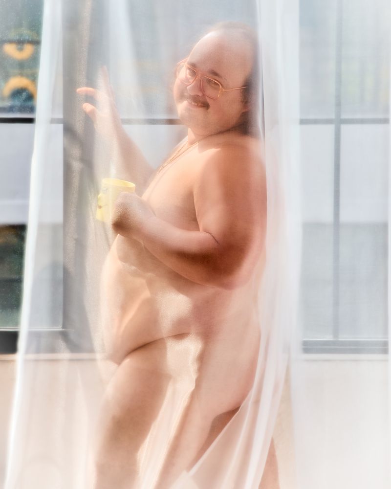 An adult man with glasses stands behind a sheer white curtain near a window, holding a yellow mug, smiling slightly, indoors.