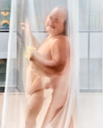 An adult man with glasses stands behind a sheer white curtain near a window, holding a yellow mug, smiling slightly, indoors.