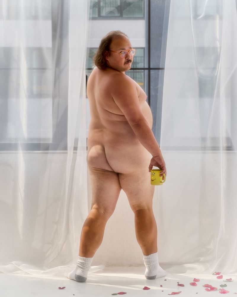 Nude adult man poses in front of window holding yellow mug, wearing white socks in indoor setting with rose petals on the floor.