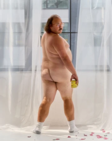 Nude adult man poses in front of window holding yellow mug, wearing white socks in indoor setting with rose petals on the floor.