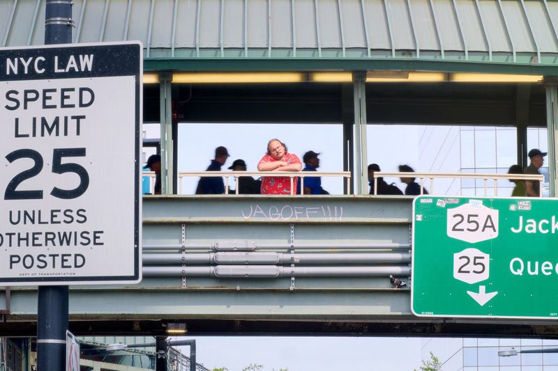 Man in red shirt and other individuals on pedestrian bridge overlooking road with traffic signs in urban setting; modern building backdrop.