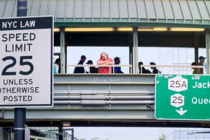 Man in red shirt and other individuals on pedestrian bridge overlooking road with traffic signs in urban setting; modern building backdrop.