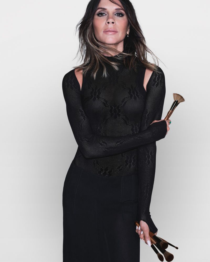 Elegant woman in black dress holds makeup brushes in a studio portrait, showcasing beauty and fashion against a plain background.