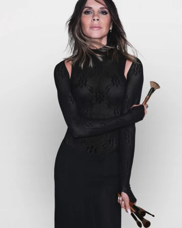 Elegant woman in black dress holds makeup brushes in a studio portrait, showcasing beauty and fashion against a plain background.