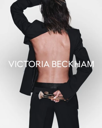 A woman with bare back wearing a black jacket and pants, holding makeup brushes, with the Victoria Beckham brand logo in the foreground.