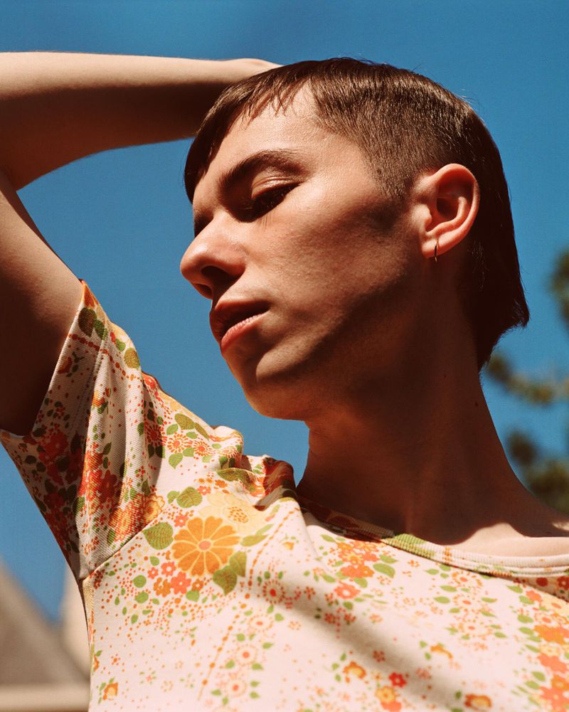 Portrait of a young adult male looking off-camera with floral-patterned top against a vibrant blue sky on a sunny day outdoors.