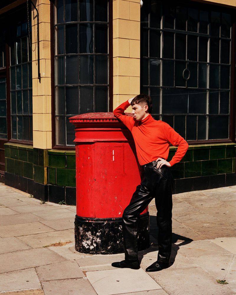 Young adult in red turtleneck leans against a red post box on a city sidewalk, wearing leather pants and black shoes near a building.