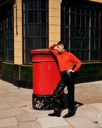 Young adult in red turtleneck leans against a red post box on a city sidewalk, wearing leather pants and black shoes near a building.