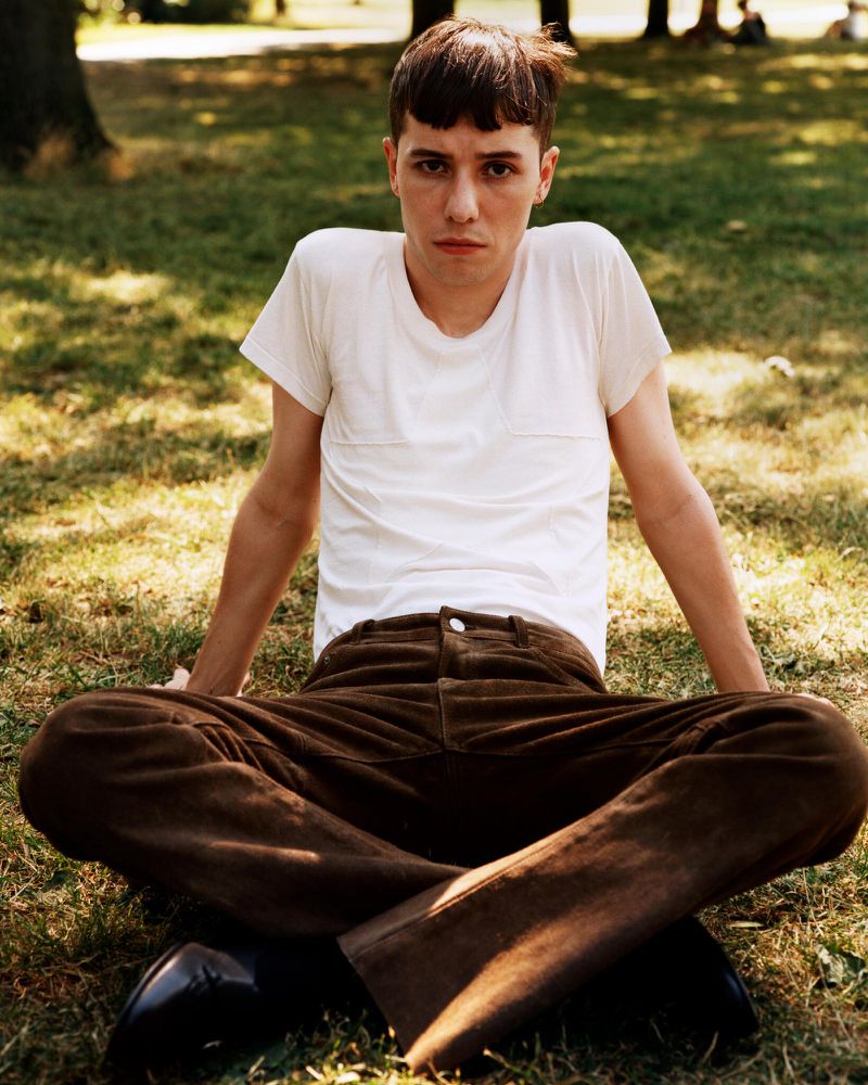 Young adult poses cross-legged on grassy lawn in casual white t-shirt and brown corduroy pants with trees in the background.
