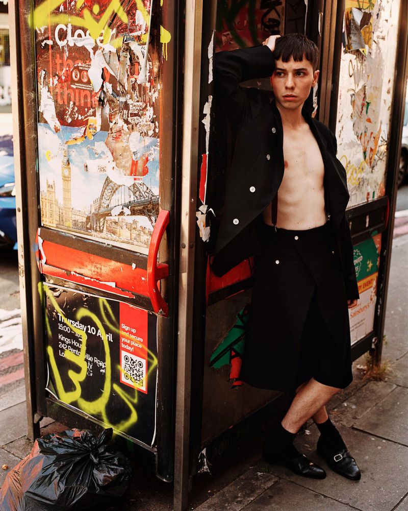 Person in stylish black attire poses inside a graffiti-covered phone booth on a city street, creating an edgy, urban fashion statement.