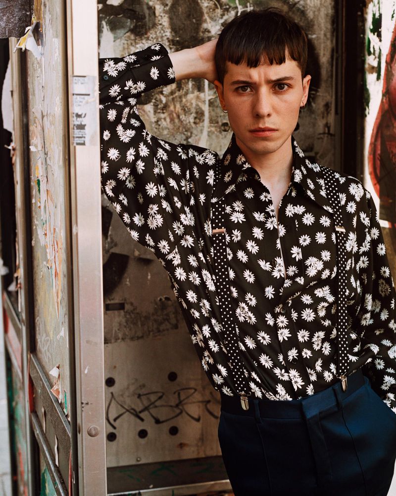 Man in daisy-print shirt and suspenders poses near graffitied wall, looking at the camera with neutral expression in medium shot.