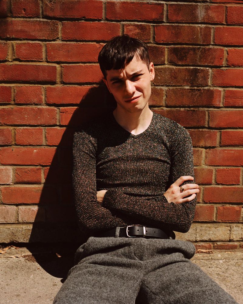 Young adult in shiny long-sleeve shirt and gray wool trousers poses against a red brick wall outdoors with sunlight and shadows.