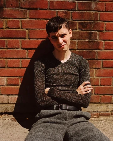 Young adult in shiny long-sleeve shirt and gray wool trousers poses against a red brick wall outdoors with sunlight and shadows.