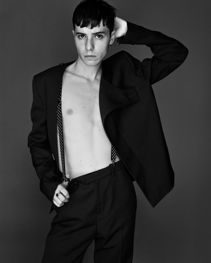 Black and white portrait of a young male adult in a black suit and suspenders posing against a gray backdrop in a photo studio.