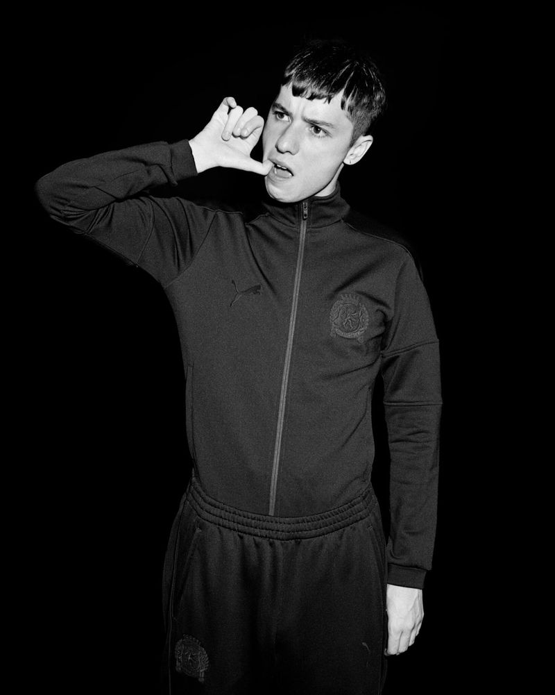 Black and white portrait of a young man in a tracksuit, posing with a hand gesture against a stark black backdrop in a studio setting.