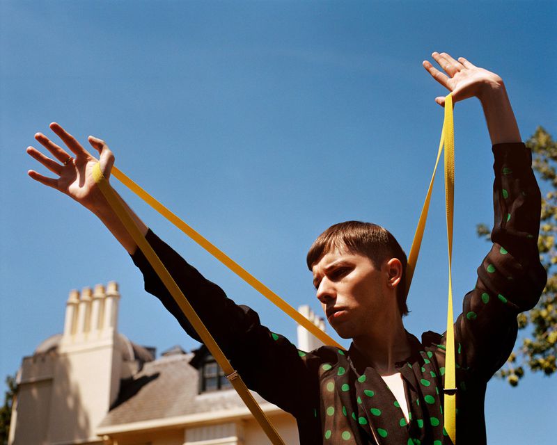 Male model poses outdoors with yellow straps against a clear blue sky, wearing a black shirt with green polka dots, building in background.
