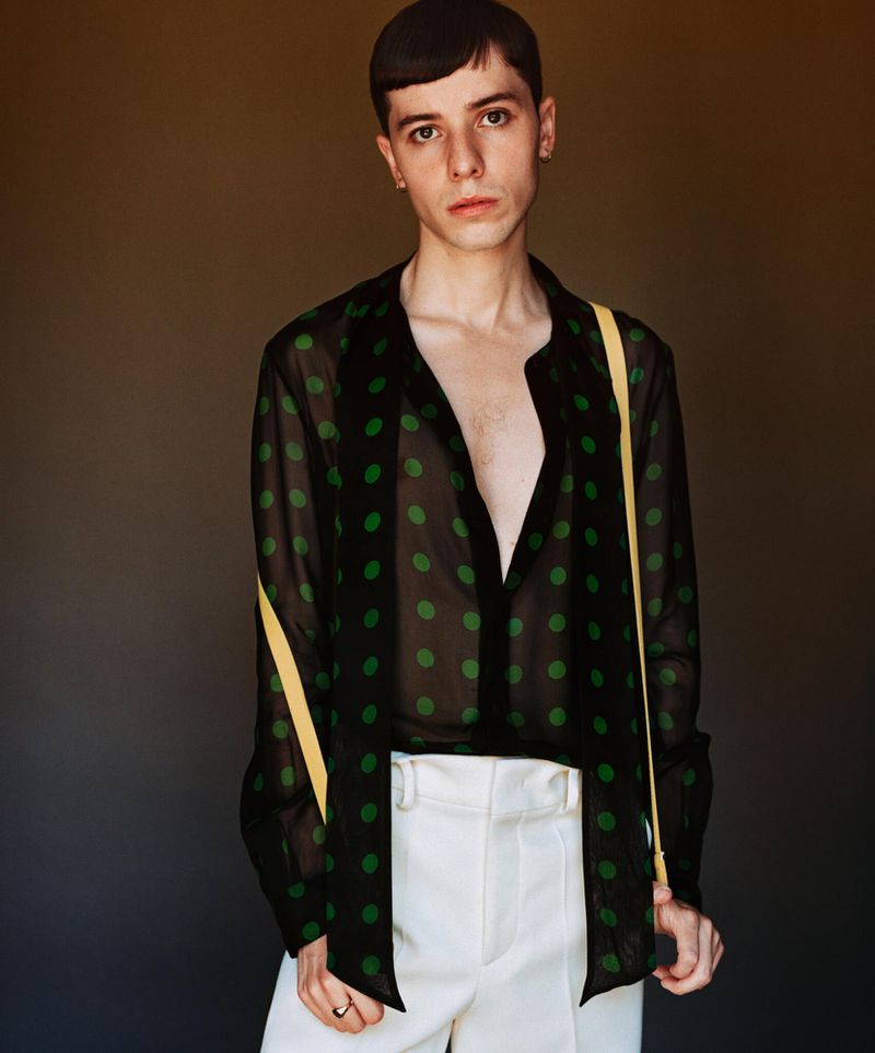 Portrait of a young adult wearing a black polka dot shirt and white pants, with a tan strap over his shoulder, indoor studio setting.