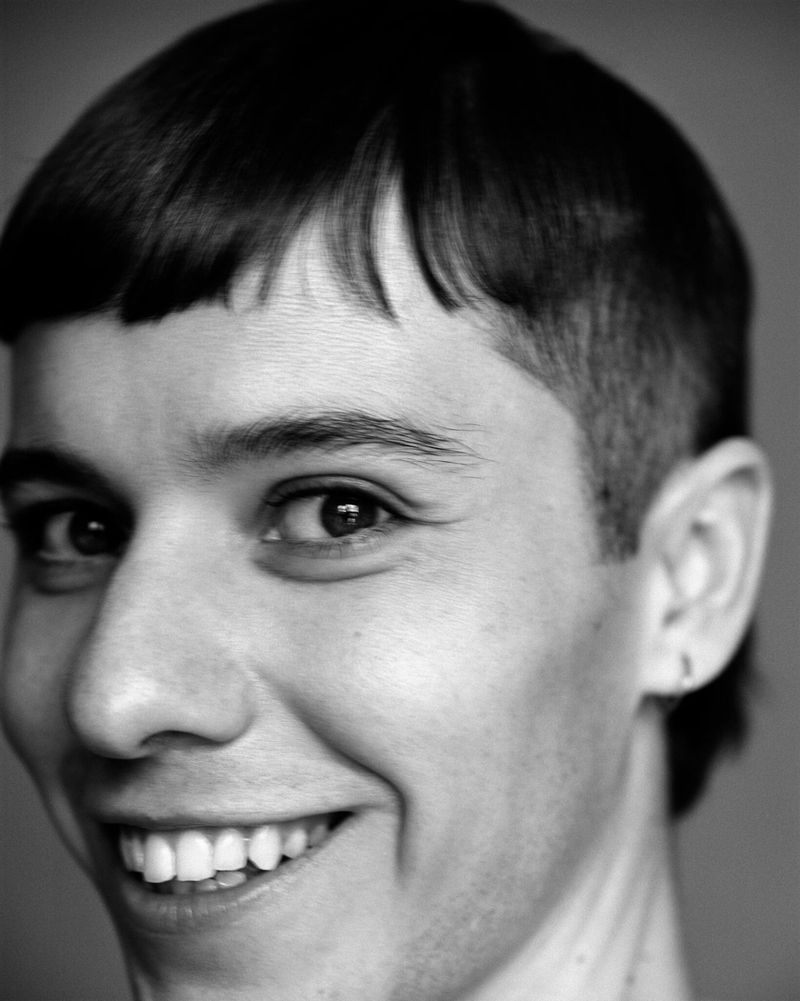 Close-up black and white portrait of a smiling adult with a bowl haircut and an earring, set against a plain gray backdrop.