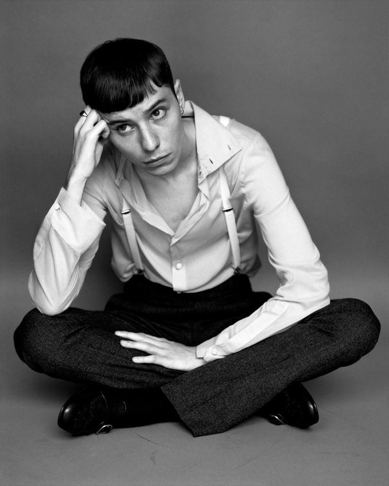 Monochrome portrait of a young adult seated cross-legged in shirt, trousers, and suspenders, against a neutral grey backdrop.