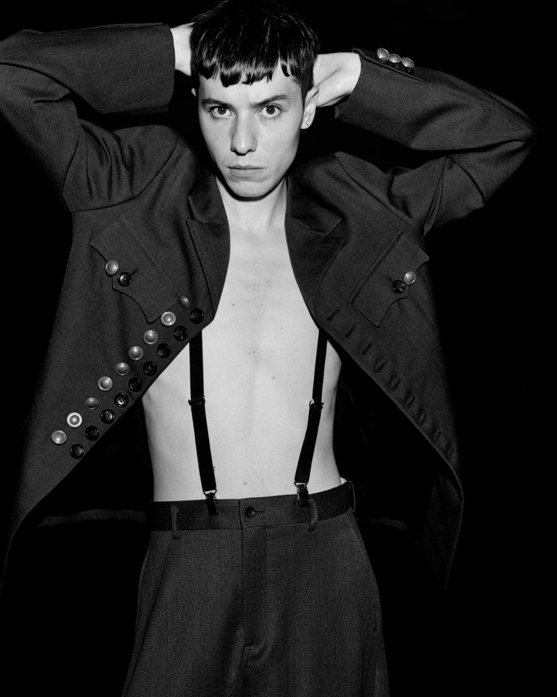 Monochrome portrait of a young man with suspenders and an open jacket, holding his hands behind his head against a black backdrop.