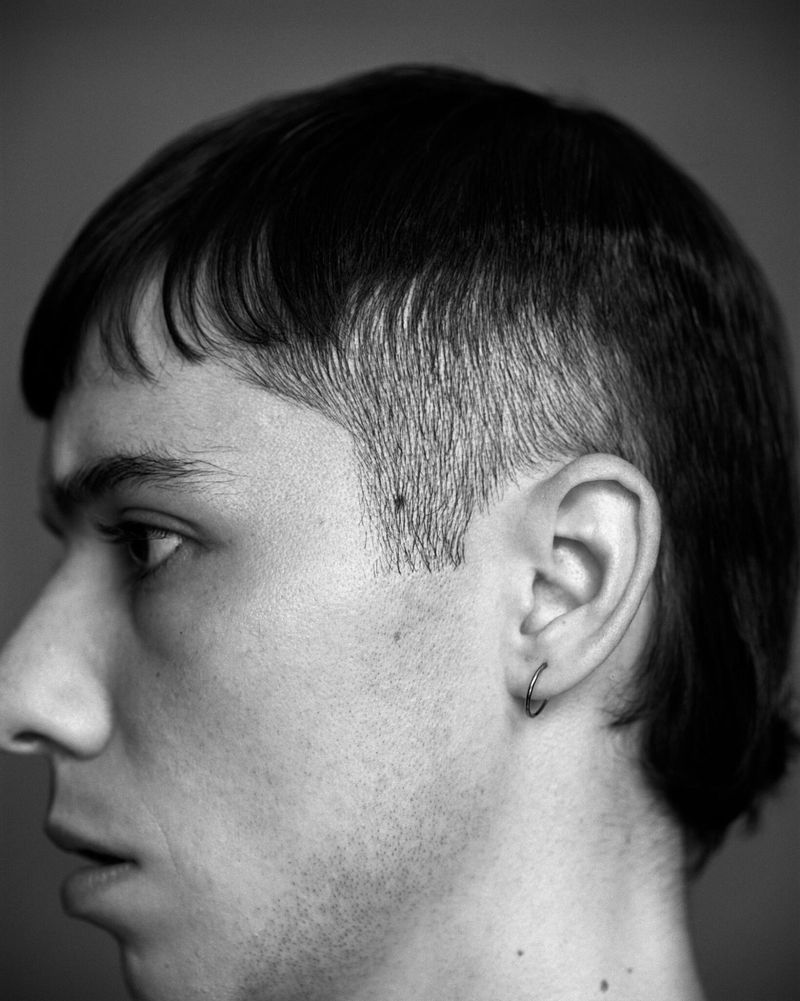 Black and white portrait of a young adult man with a mullet hairstyle, captured in a profile shot against a neutral background.