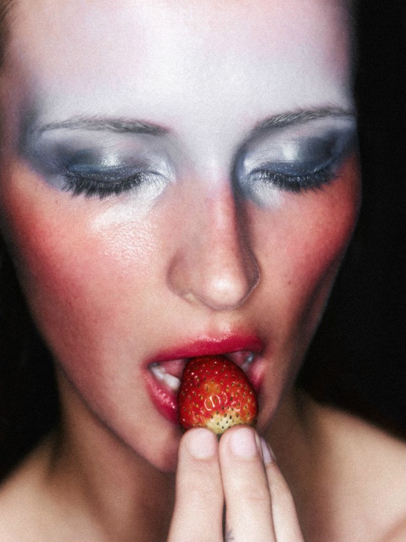 Close-up of a person with dramatic makeup holding a strawberry to their mouth, set against a dark background for a beauty-focused image.