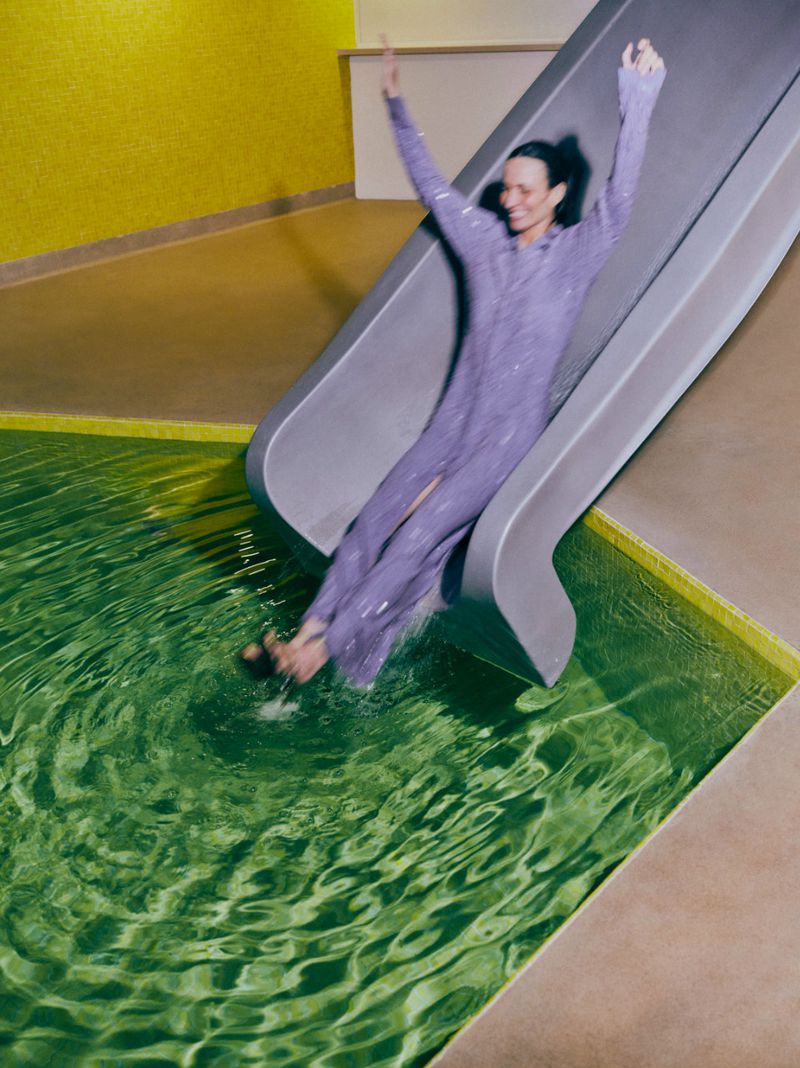 Smiling woman in a sequined dress sliding into a green-tiled indoor pool with arms raised, set against a yellow tiled wall.