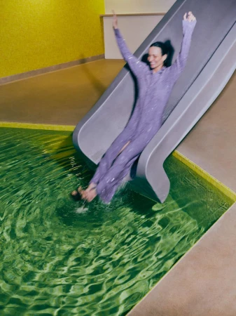 Smiling woman in a sequined dress sliding into a green-tiled indoor pool with arms raised, set against a yellow tiled wall.