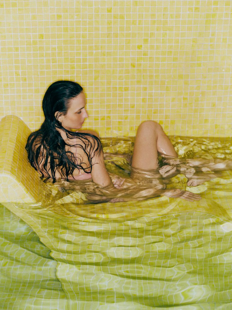 Woman relaxing in a bathtub filled with clear water, surrounded by yellow mosaic tiles, creating a serene spa-like atmosphere.