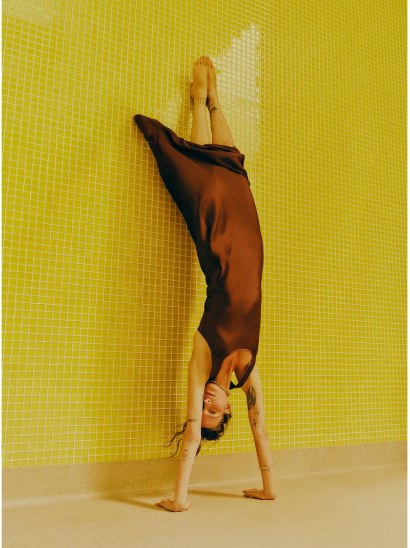 Adult woman in a brown dress doing a handstand against a yellow tiled wall, demonstrating fitness and body-positive lifestyle.