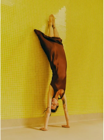 Adult woman in a brown dress doing a handstand against a yellow tiled wall, demonstrating fitness and body-positive lifestyle.