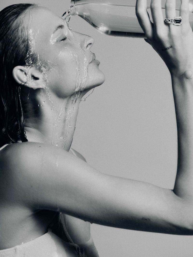 Monochrome portrait of a woman with wet hair and closed eyes, water pouring on her face from a clear jug, against a neutral backdrop.
