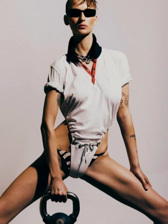 Woman in sunglasses poses with a kettlebell wearing a white polo shirt and striped bikini bottoms against a light background in studio.