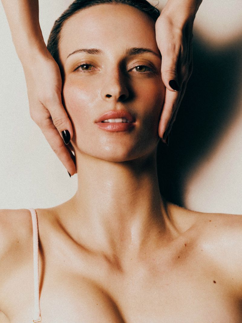 Close-up studio shot of a fair-skinned woman receiving a facial massage with hands visible and light background.