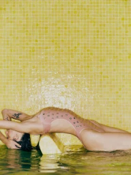 Model wearing a pink one-piece swimsuit lounging in water against a yellow tiled wall for Vogue Brasil by Hick Duarte