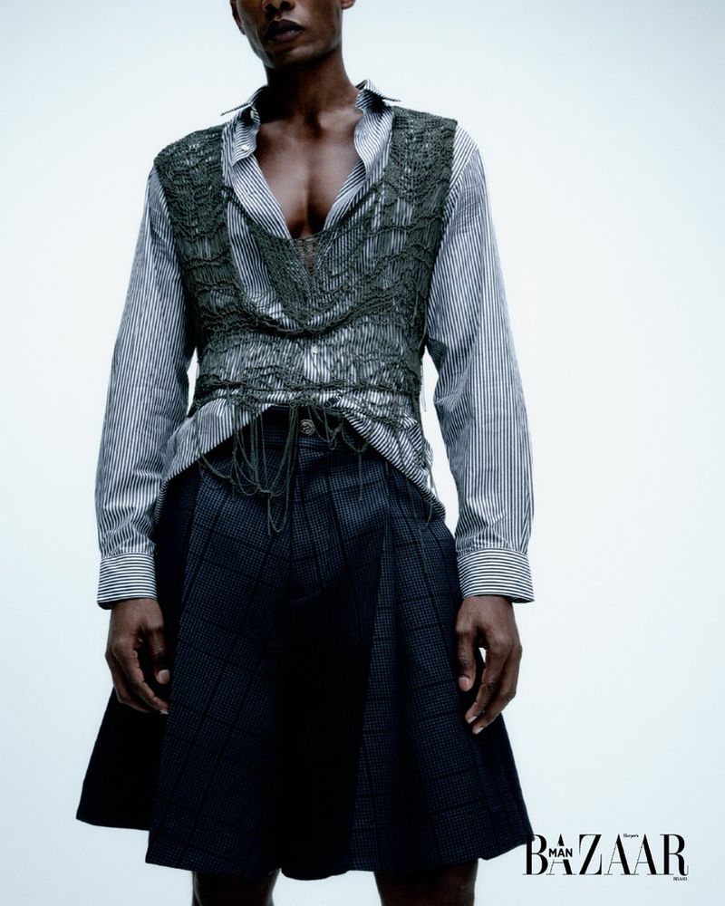 Dark-skinned adult male fashion model poses in a striped shirt, decorative vest, and plaid skirt-shorts in a studio setting.