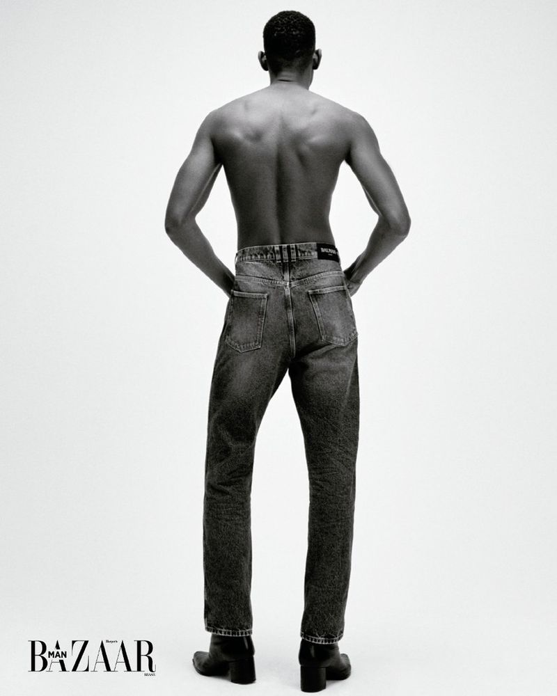 Back view of a shirtless African American man wearing dark denim jeans and black boots against a plain white background in a studio shot.