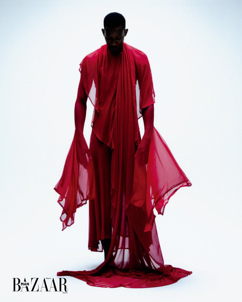 A striking fashion shot of a Black male model in red fabric posing in a photo studio wearing draped chiffon. Part of a Man Bazaar editorial.