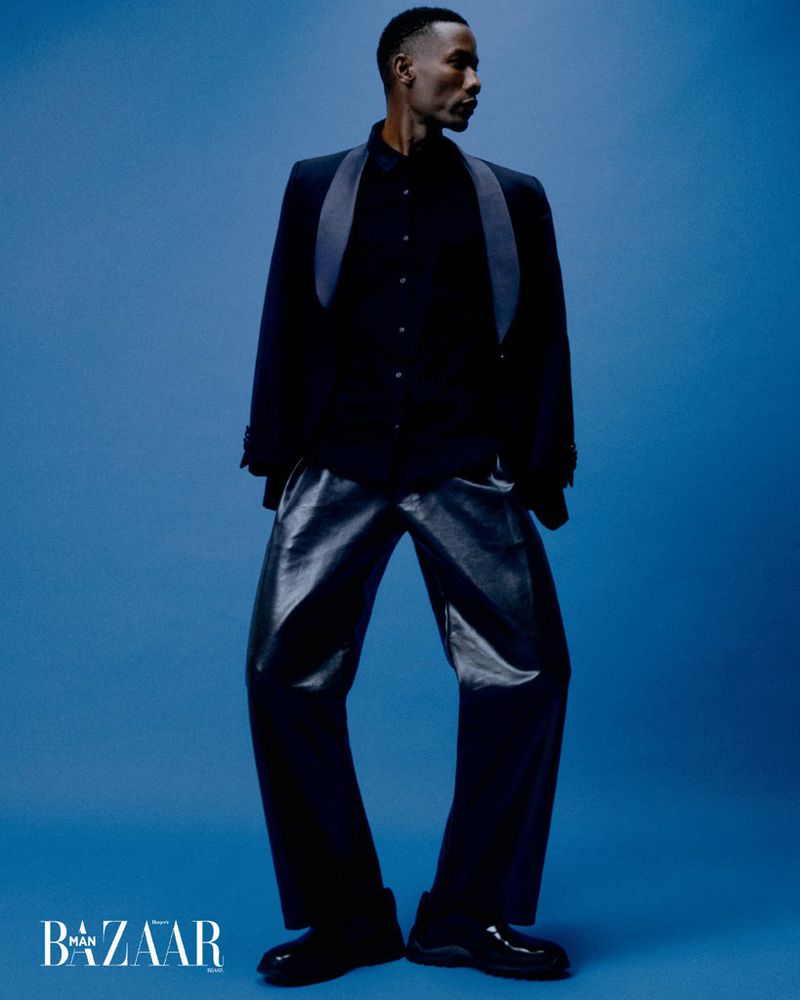 Full-length studio shot of a dark-skinned male fashion model in a dark suit and leather pants against a gradient blue backdrop.