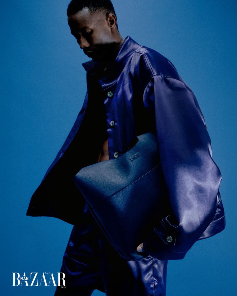 Dark-skinned man in bright blue satin outfit with bag, fashion-style studio shot, standing against a matching blue backdrop with logo.
