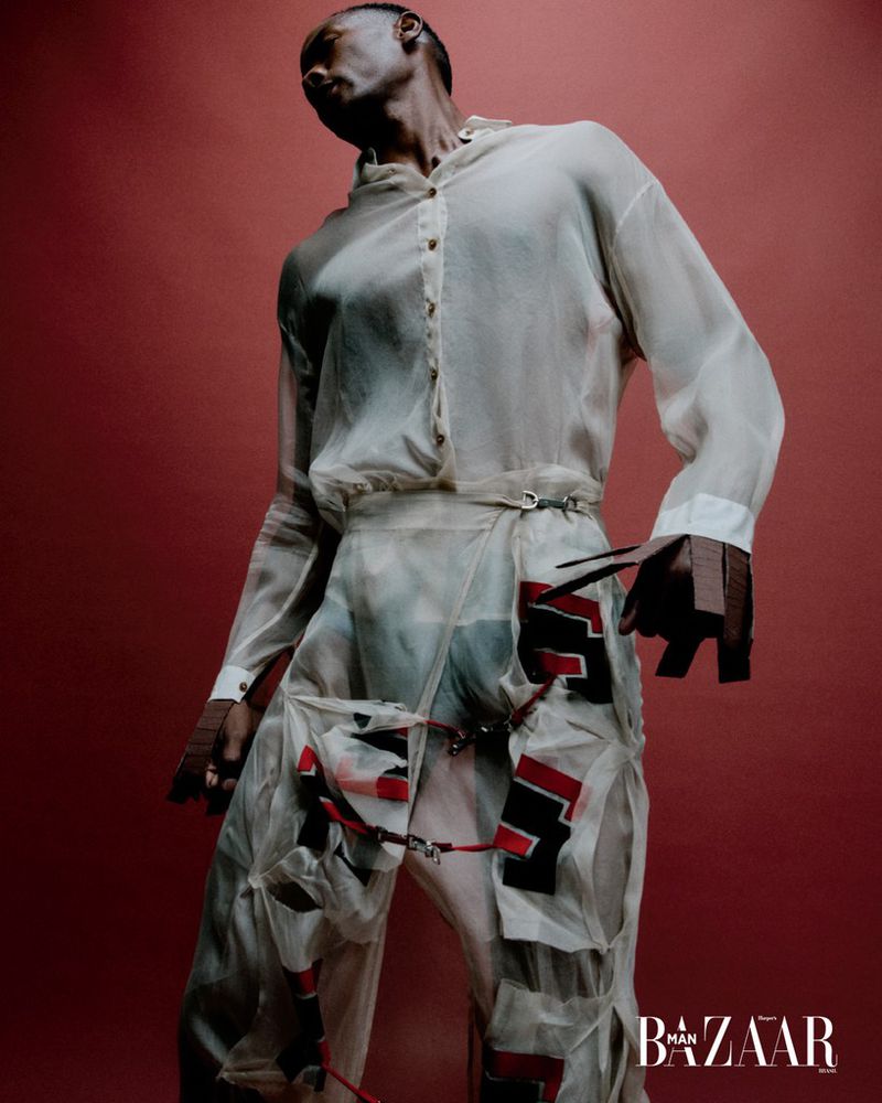 Fashion editorial shot of a Black male model in translucent white shirt and patchwork pants with a red background and Bazaar logo.