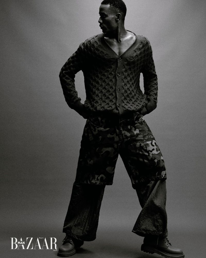 Monochrome fashion shot of an adult male model in a buttoned knit cardigan, patterned pants, and boots in a studio setting.