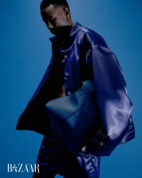 Black man wearing blue satin jacket and matching trousers holding dark handbag against solid blue background