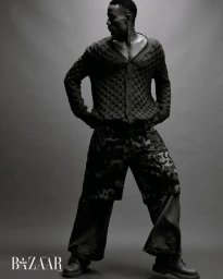 Black man wearing textured cardigan and patterned trousers standing against a plain seamless grey background.