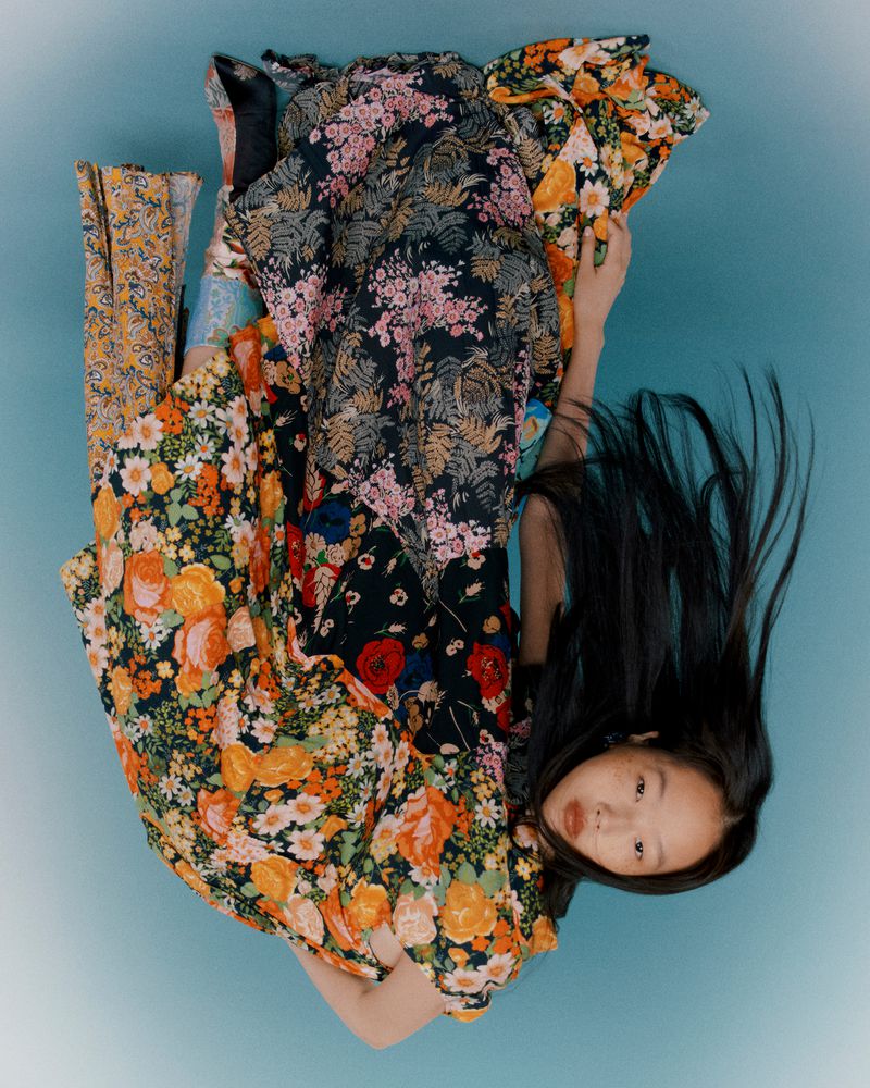 An Asian woman lies in a blue studio surrounded by multiple floral dresses and pants, showcasing a vibrant fashion-forward scene.
