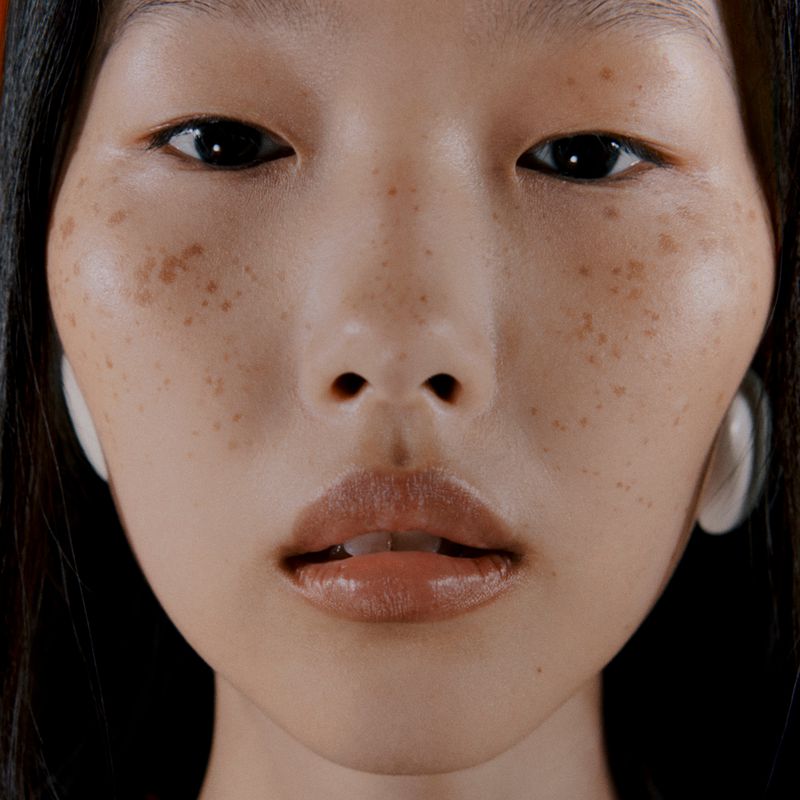 Close-up portrait of an Asian female with dark hair, freckles, and wearing large, round, light-colored earrings against a dark orange backdrop.